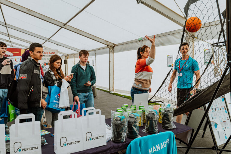 University students at a freshers fair stall trying to score a basketball in a hoop to win a free PureGym prize.