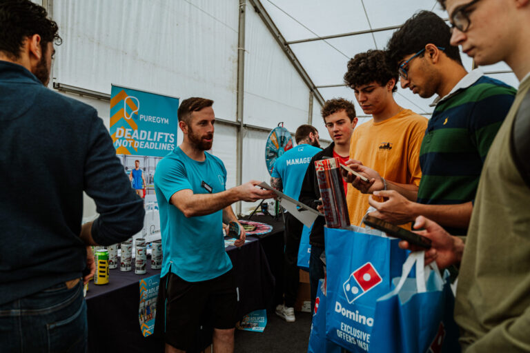 PureGym staff member at a freshers fair, handing out leaflets to a group of students.